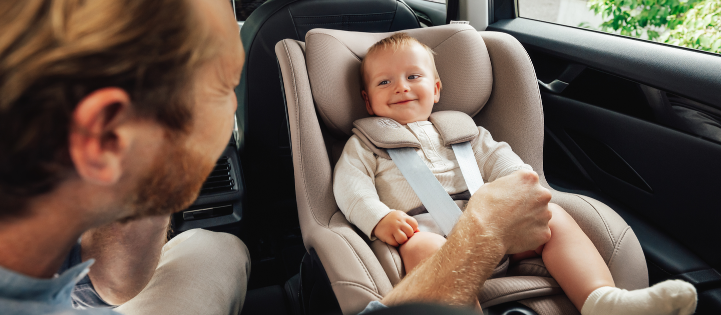 A smiling baby sits securely in a Britax R&ouml;mer Dualfix 5Z car seat in the color Teak, mounted in the back seat of a car. An adult, seen from the side, gently holds the baby&rsquo;s leg while interacting with them.